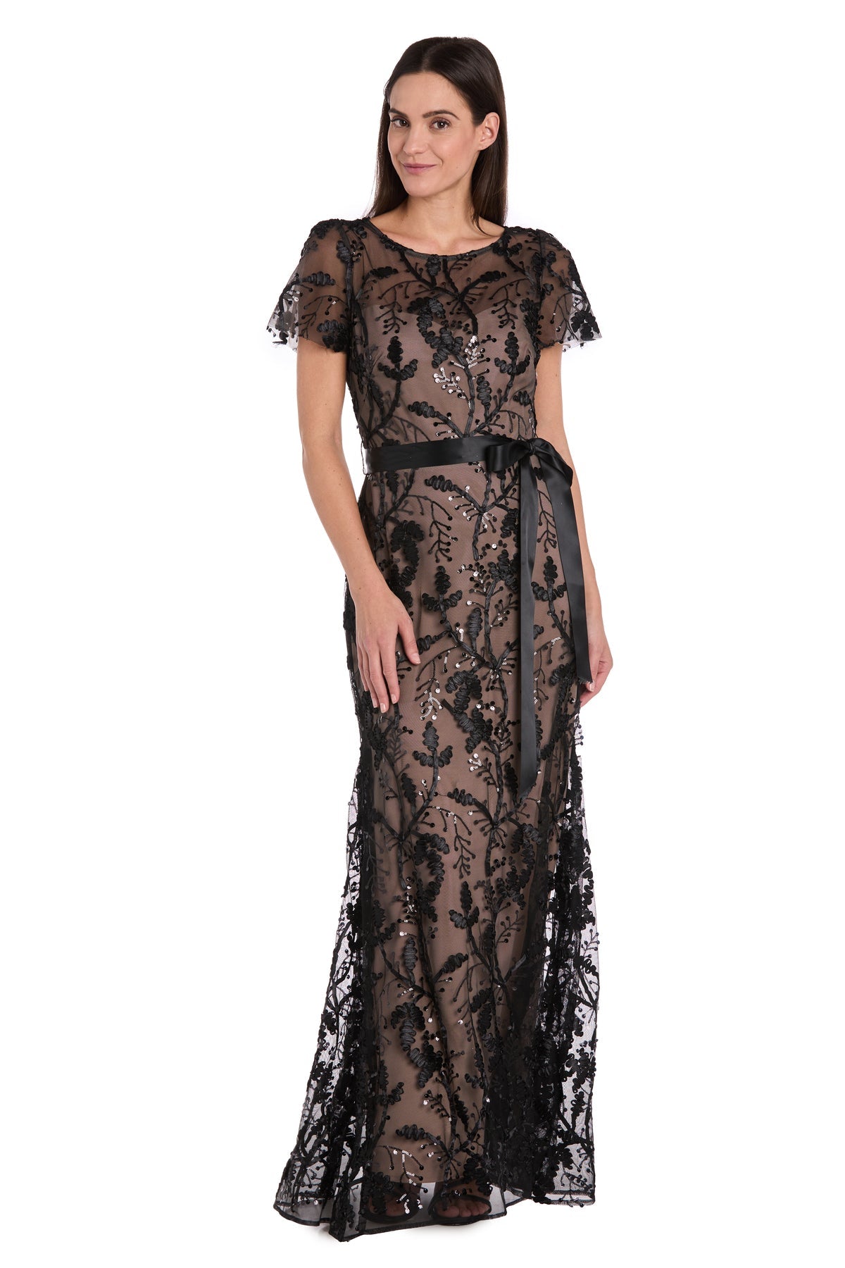 A woman in the R&M Richards 3253 short sleeve black formal dress with lace floral patterns and a satin belt stands against a white background.